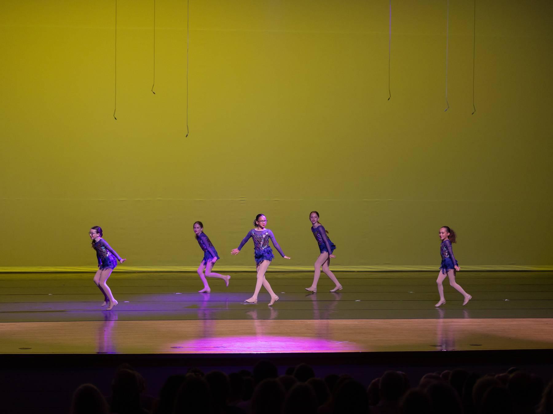 Dancers in purple costumes spread across a yellow-lit stage