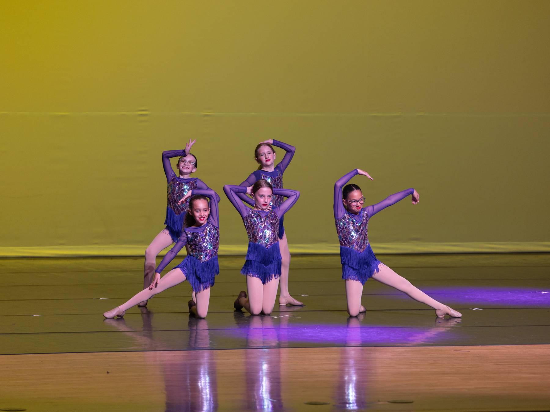 Five dancers in purple sequined costumes posed together on a yellow-lit stage