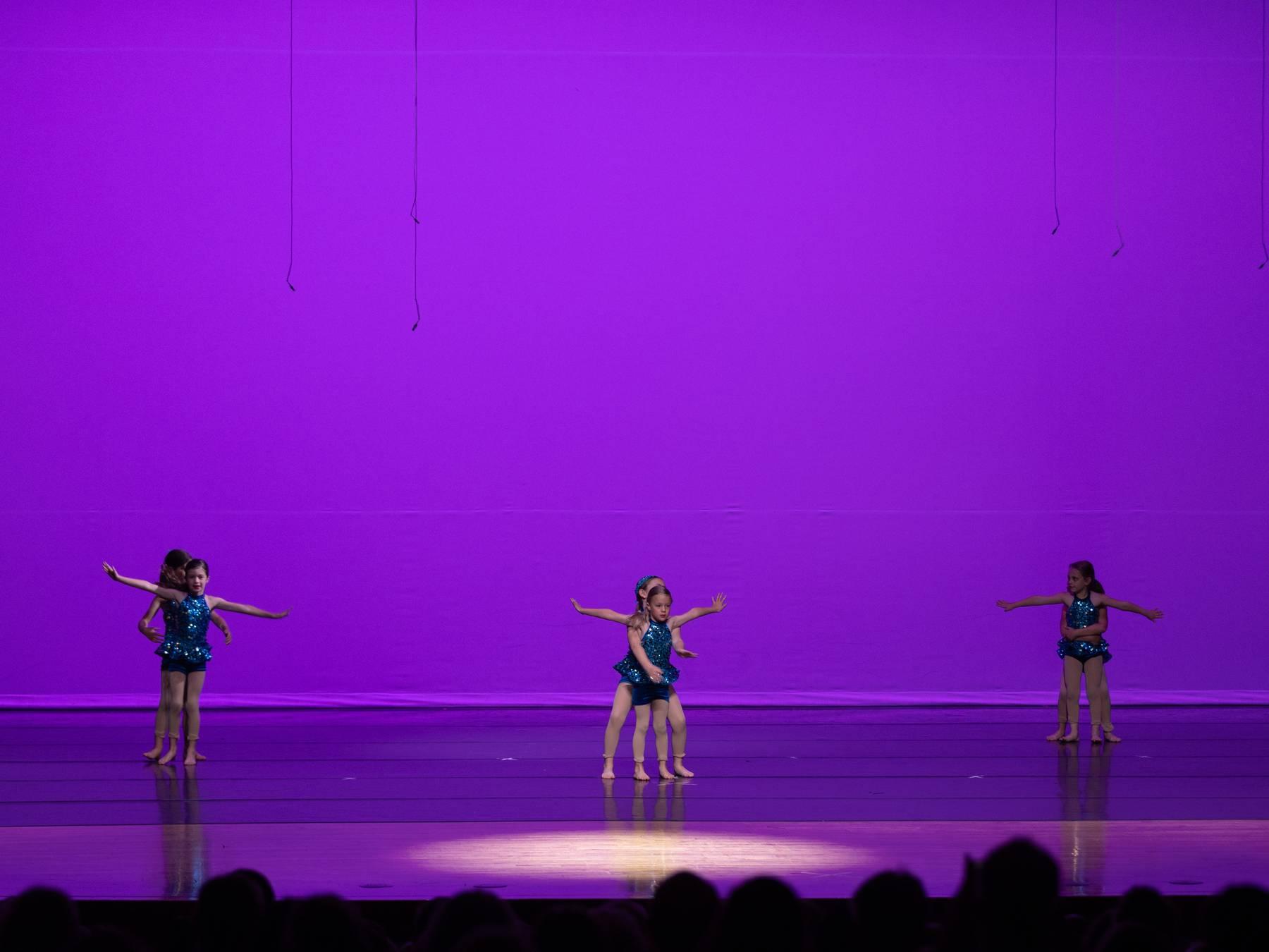 Three dancers in teal costumes with arms outstretched on a purple-lit stage
