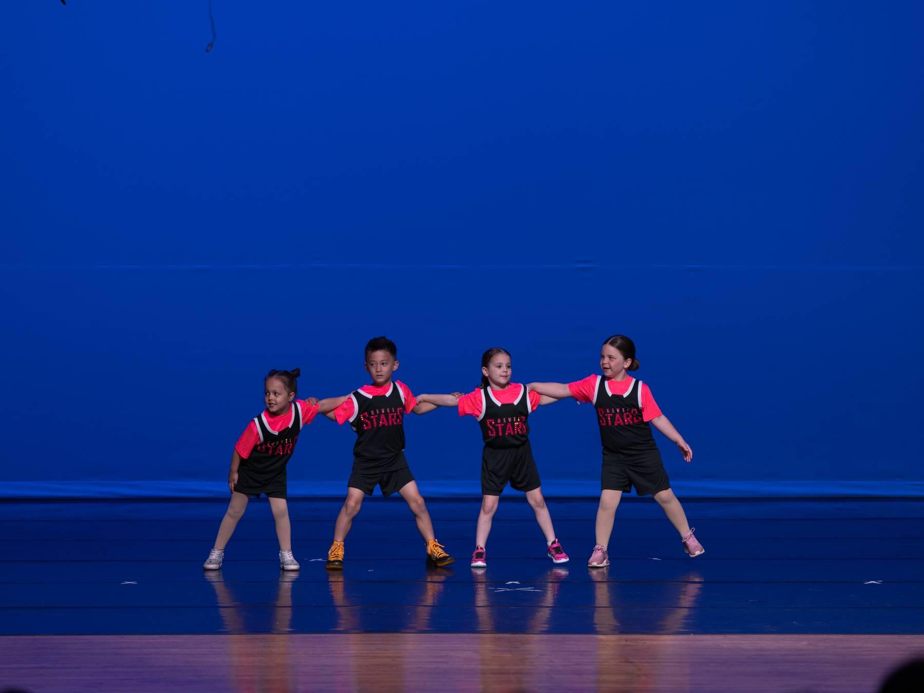 Four dancers in black and red jerseys with arms linked on a blue-lit stage