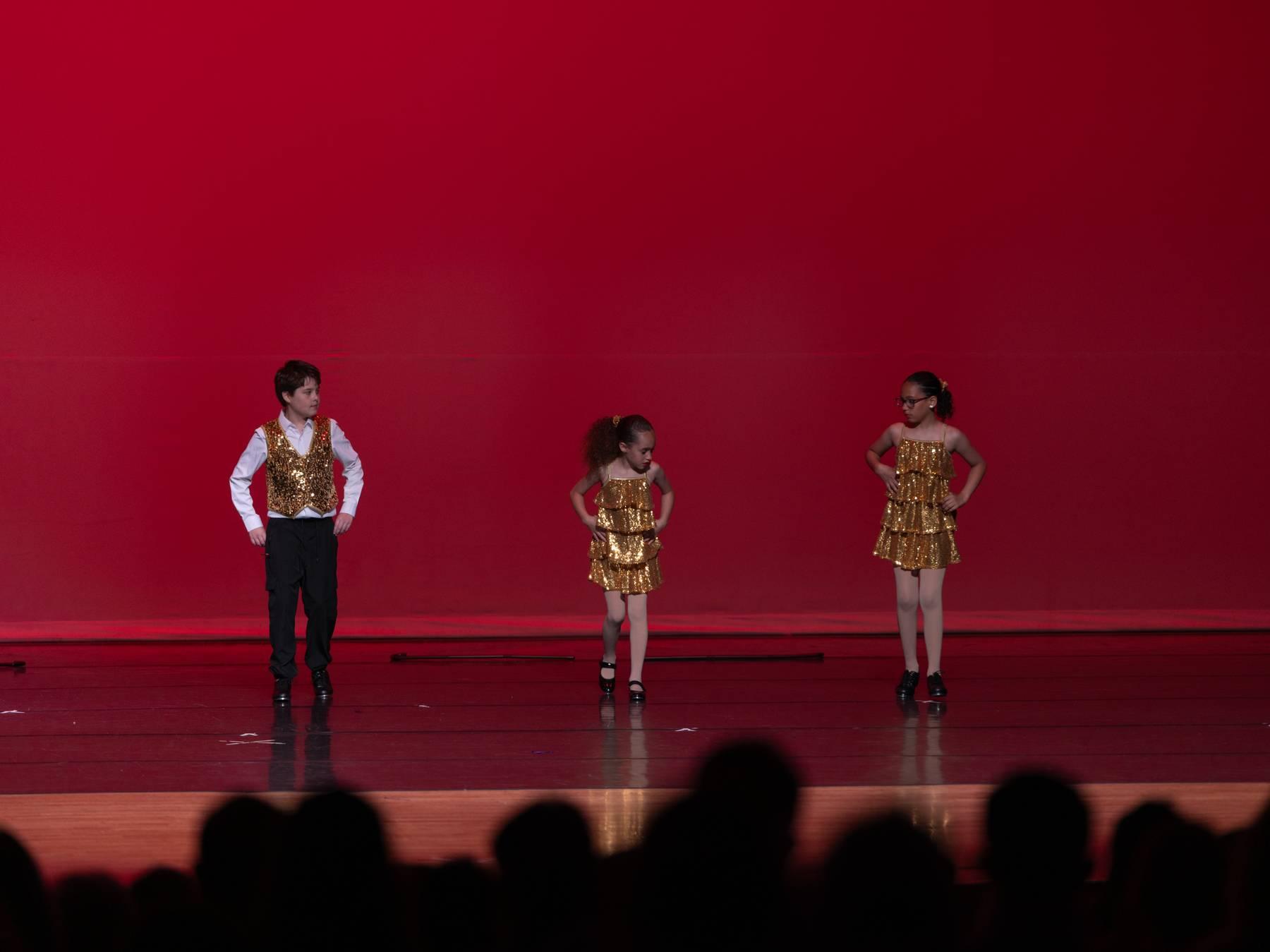 A dancer in a gold vest and two dancers in gold fringe on a red-lit stage