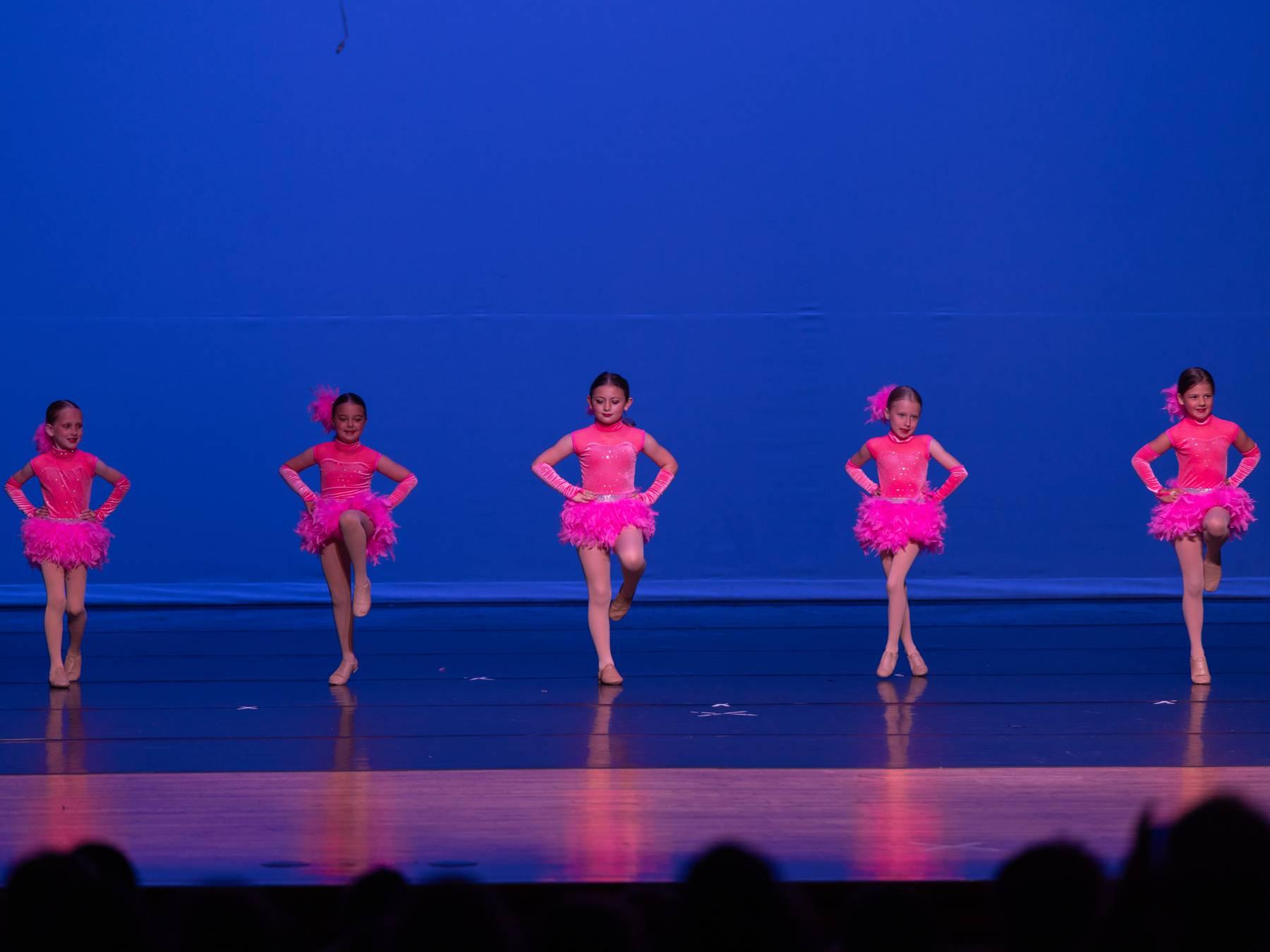 Five dancers in pink feathered costumes in a row on a blue-lit stage