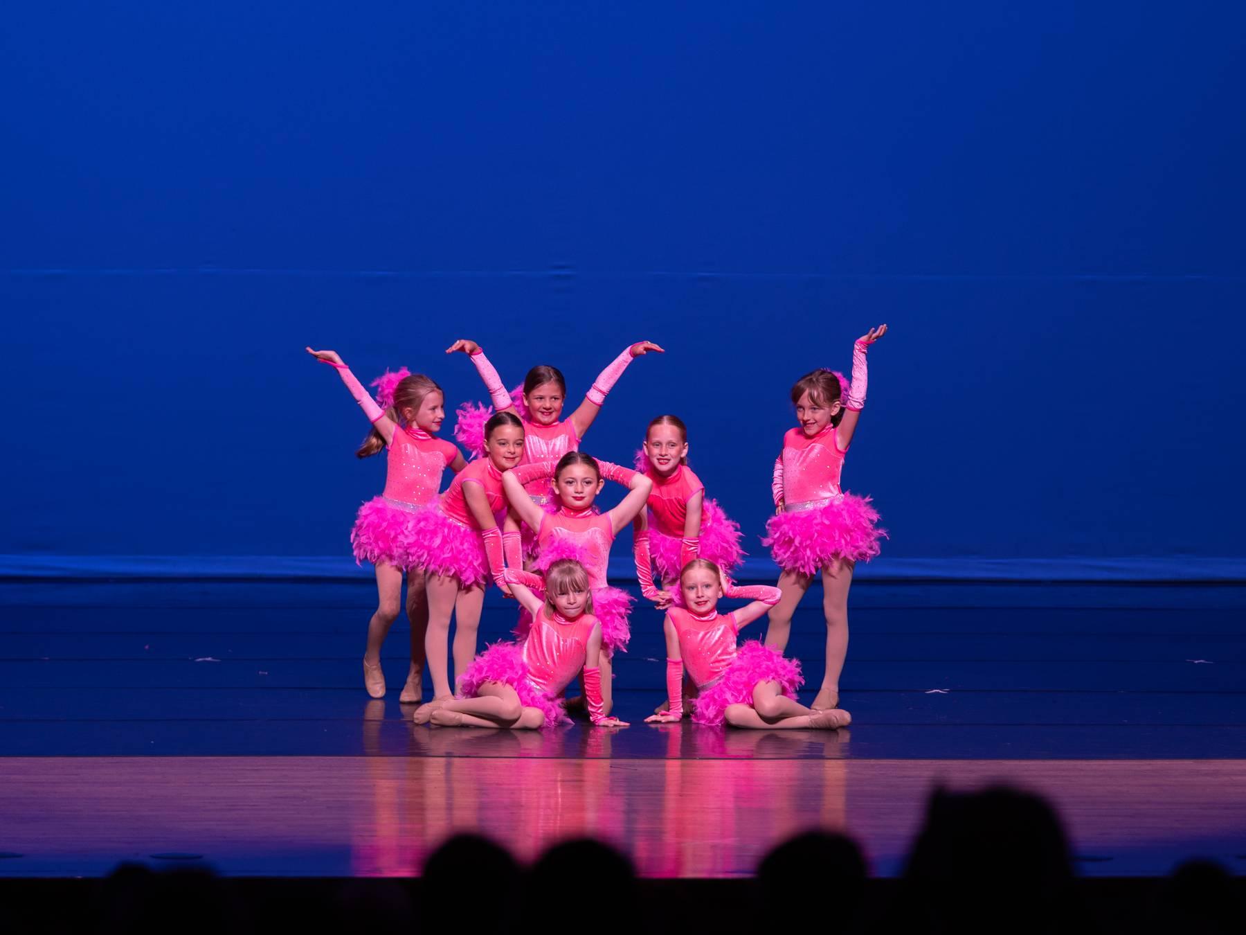 Young dancers in pink feathers posed together on a blue-lit stage