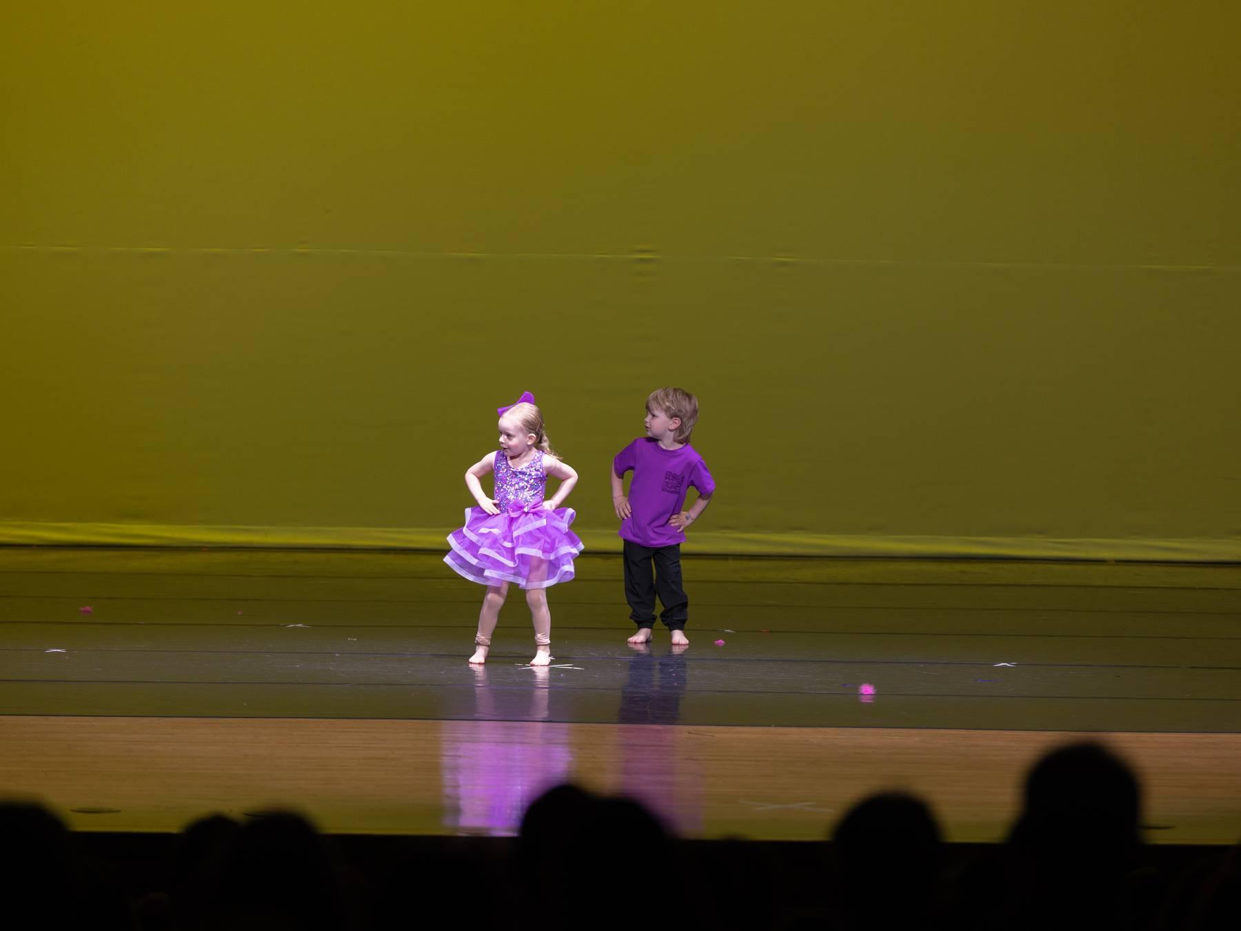 Two of the youngest dancers, one in a purple tutu, on a yellow-lit stage