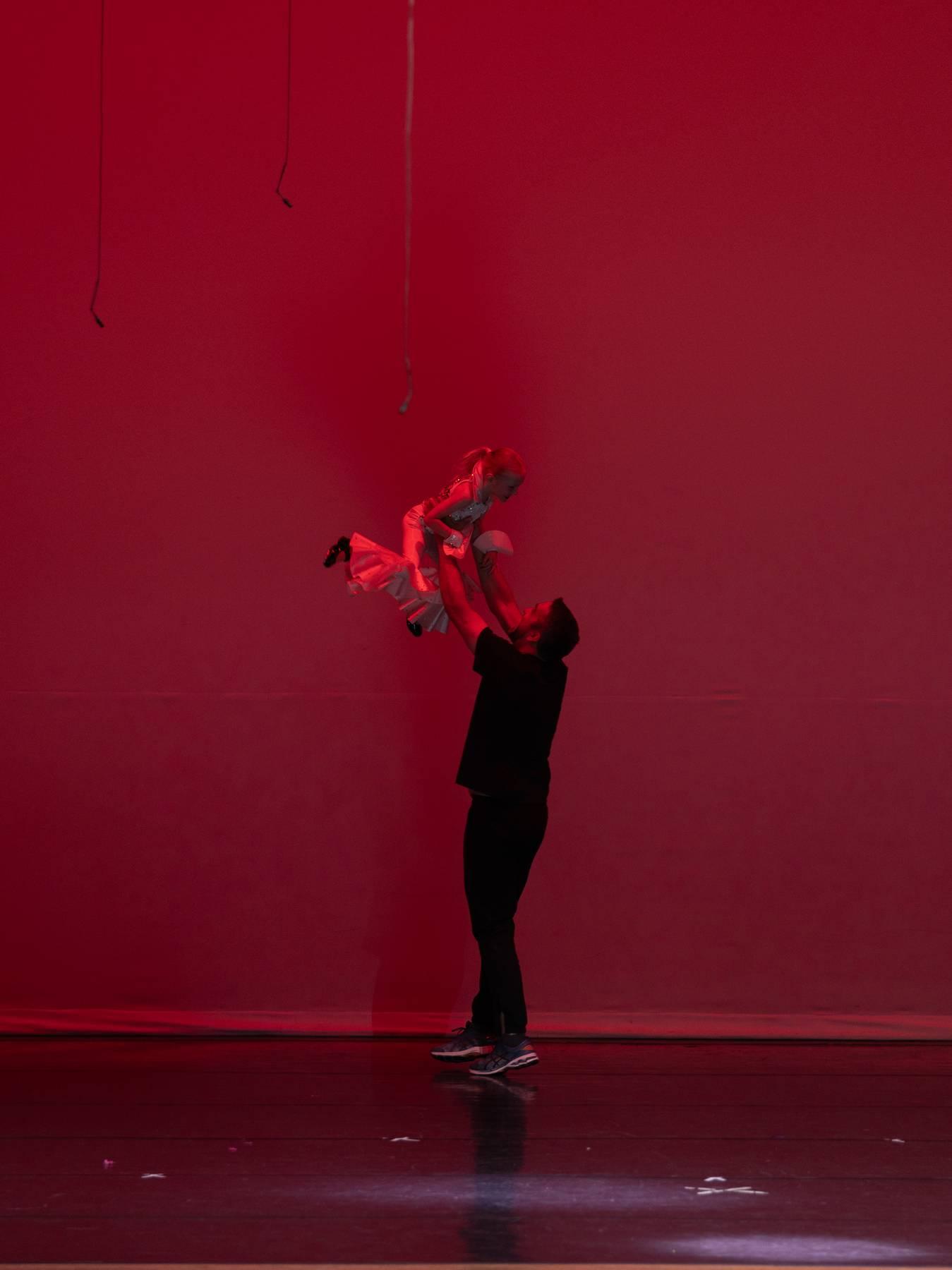 A parent lifting a young dancer overhead in front of a red backdrop