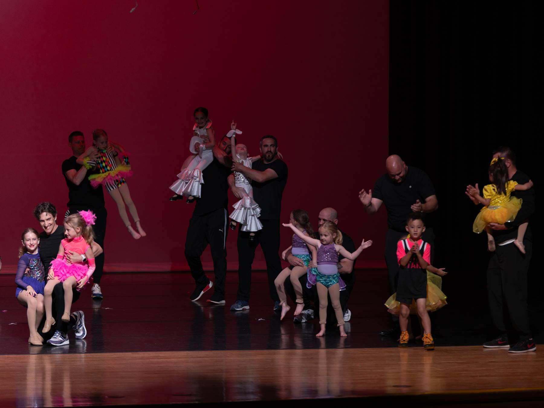 Parents holding and dancing with young dancers on a red-lit stage