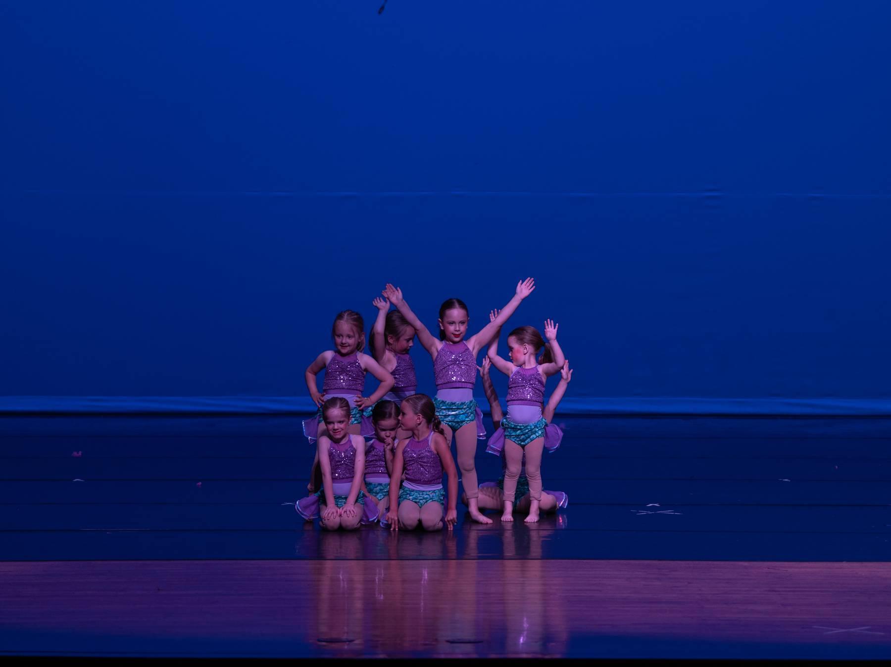 A small group of young dancers in purple and teal posed on a blue-lit stage