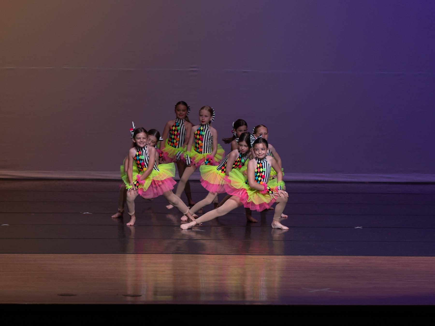 Dancers in neon green and pink tutus posed in a cluster on a purple-lit stage