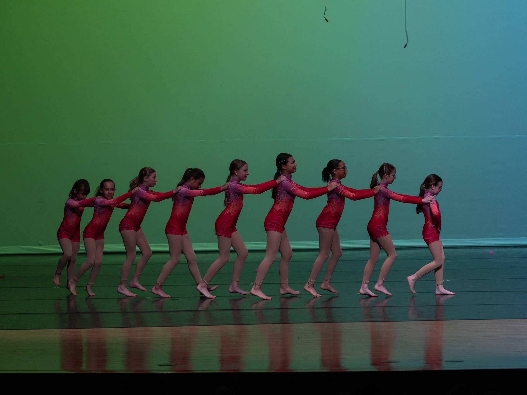 A line of dancers in red leotards linked in a chain on a green-lit stage