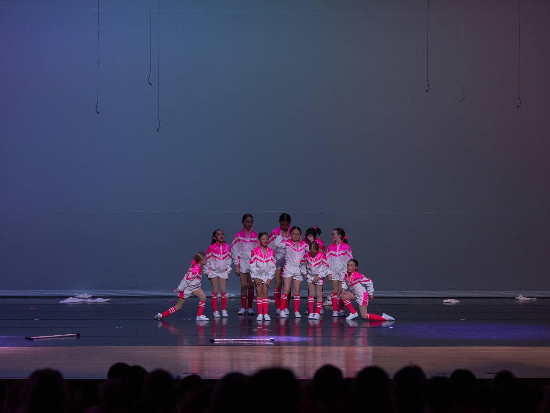 A group of dancers in pink and white jackets posed on a blue-grey stage
